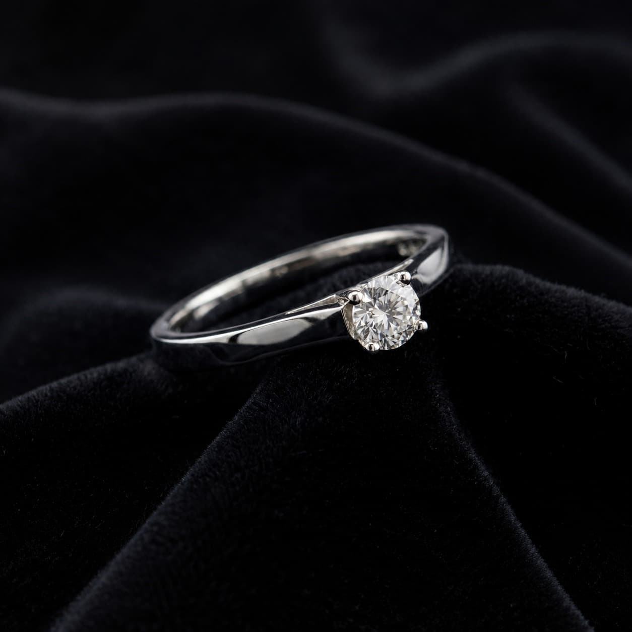 Ein funkelnder Solitär-Diamantring in Krappenfassung auf einem eleganten, tiefschwarzen Hintergrund aus weichem Samt.