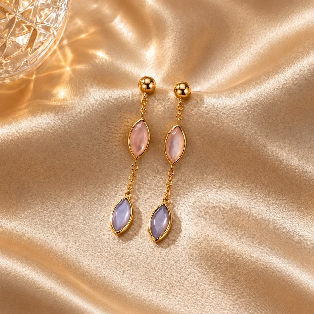 Goldene Hängeohrringe mit rosa und lila marquiseförmigen Steinen auf glänzendem, goldenem Satinstoff drapiert.